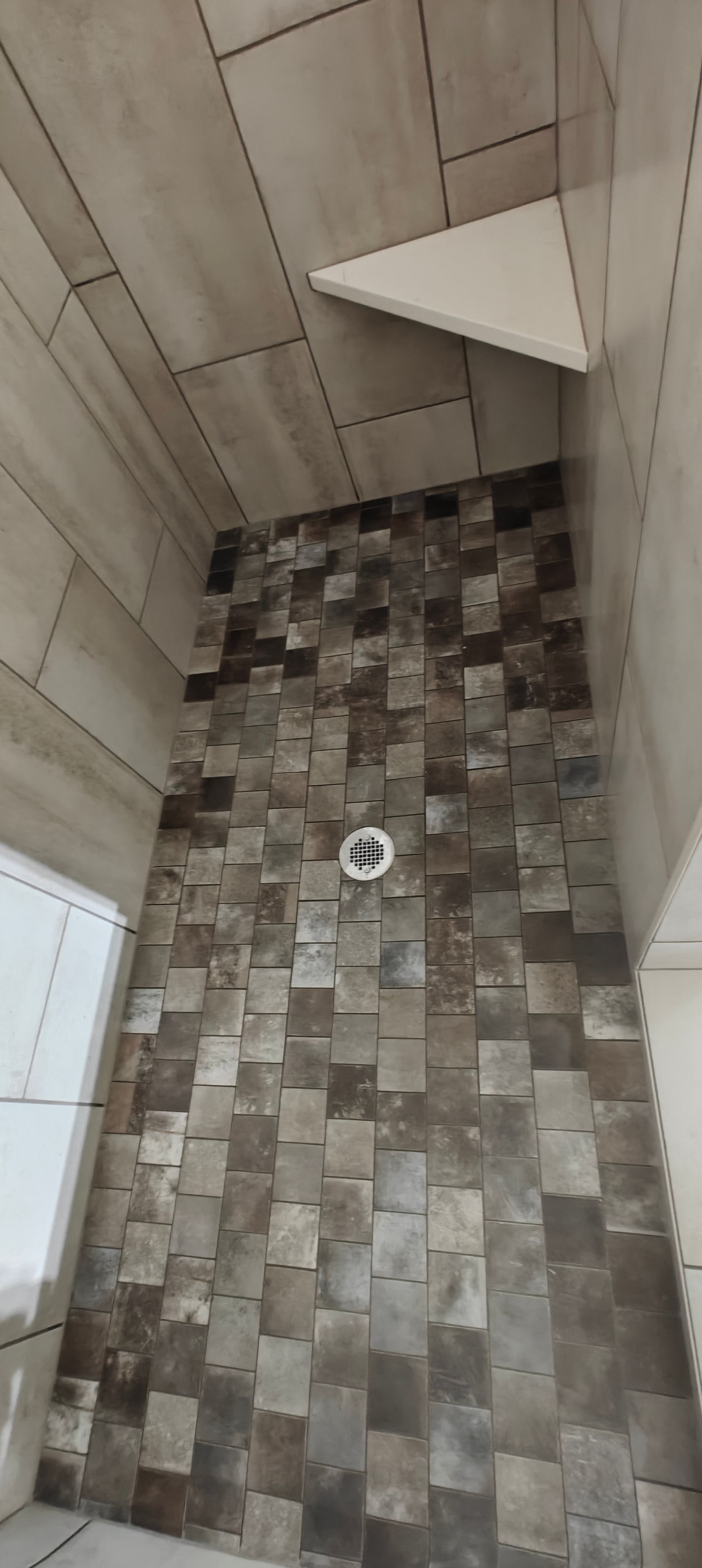 Small tiled bathroom with a triangular shower shelf and a floor drain beneath a slanted wood-paneled ceiling.