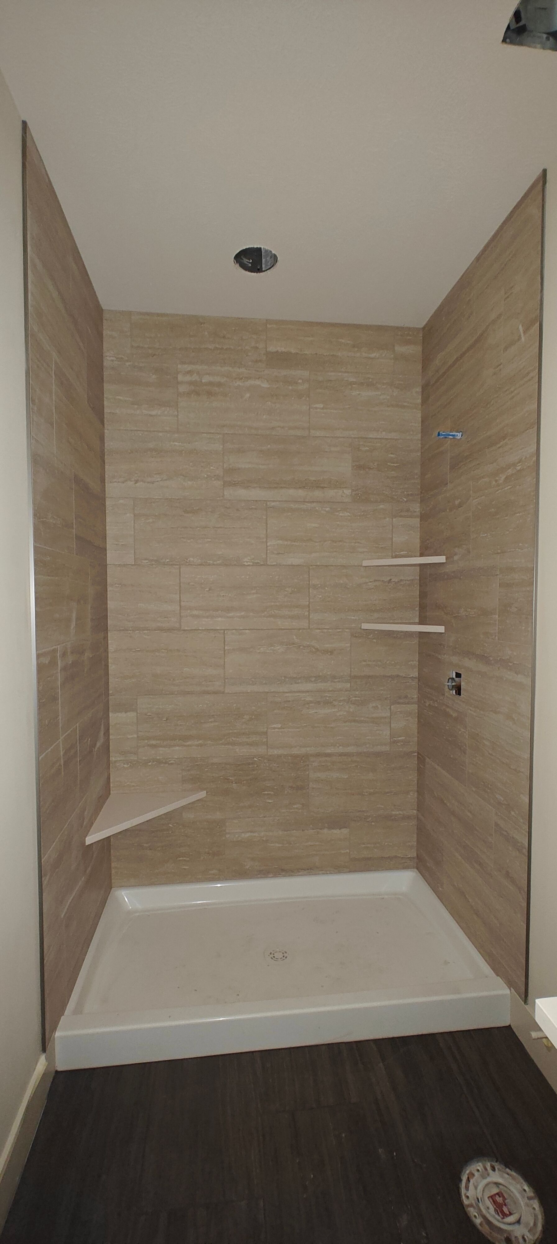 Small tiled walk-in shower with white base, beige walls, and dark wood flooring outside.