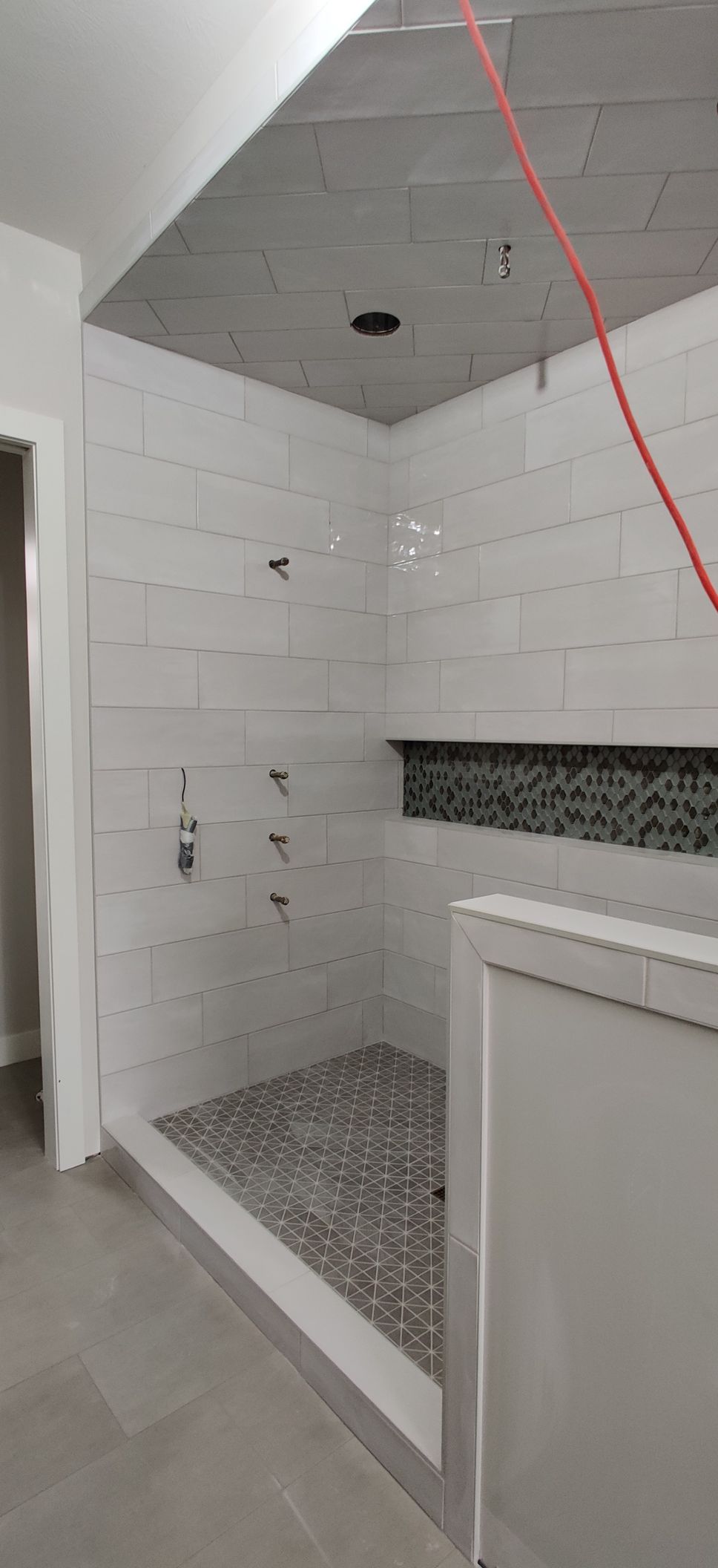 Partially tiled bathroom shower with gray ceiling, white walls, and mosaic floor tiles.