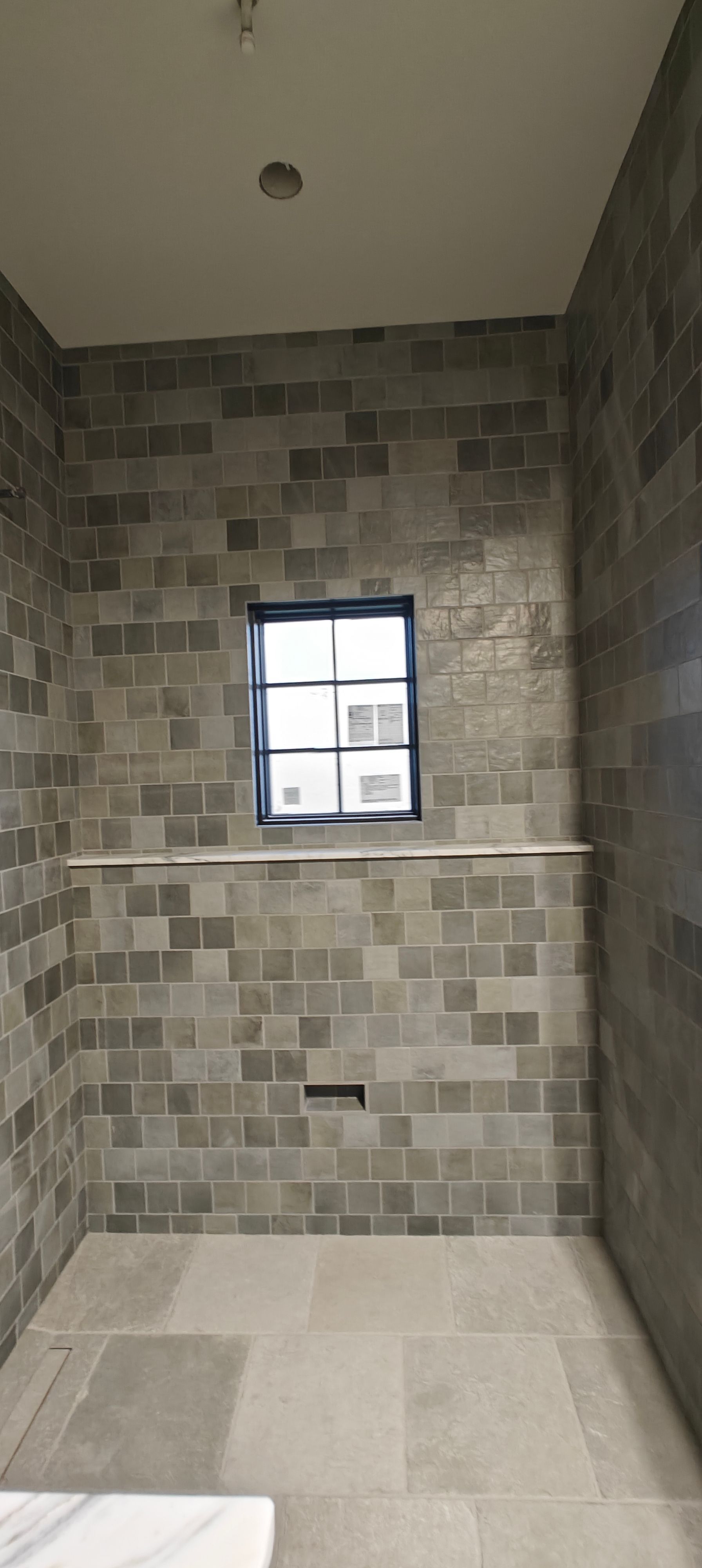 Small tiled shower stall with a window and light stone floor.