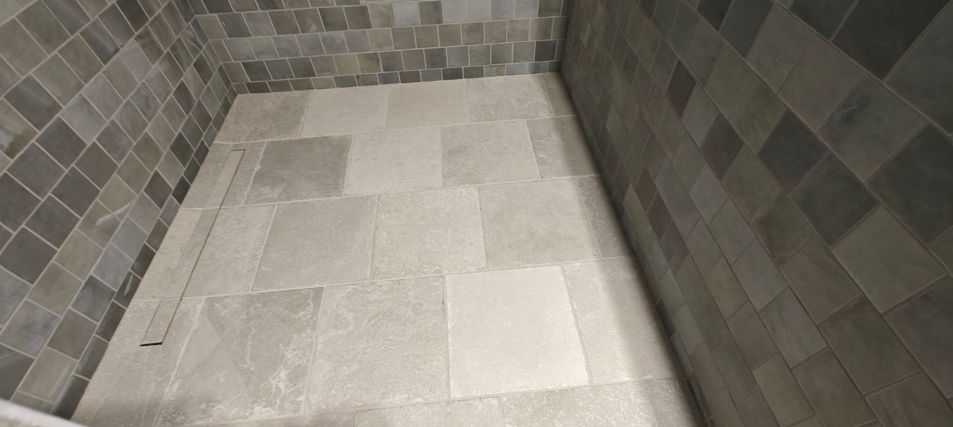 Tiled shower floor with gray mosaic walls and light stone floor tiles.