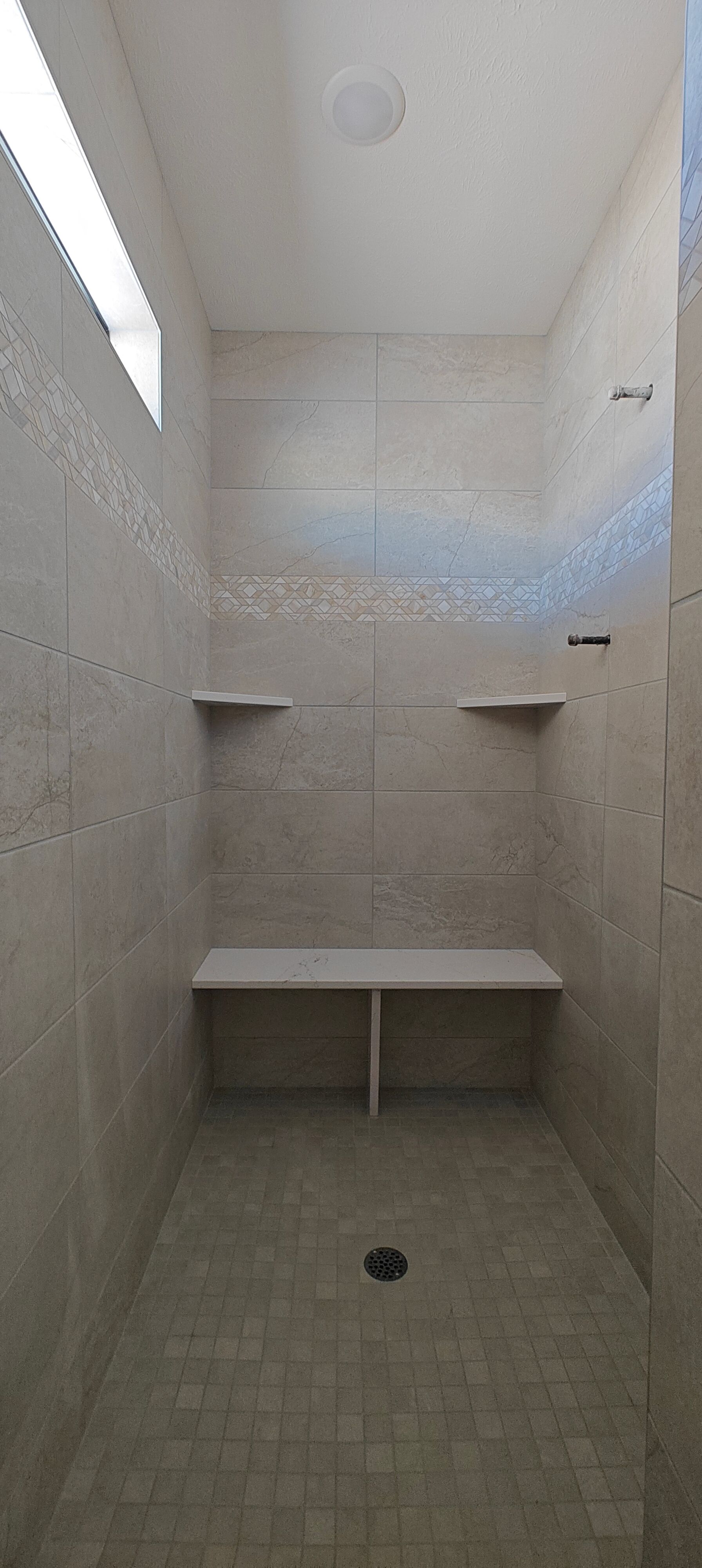 Empty tiled shower with built-in bench, shelves, and a floor drain under a small window.