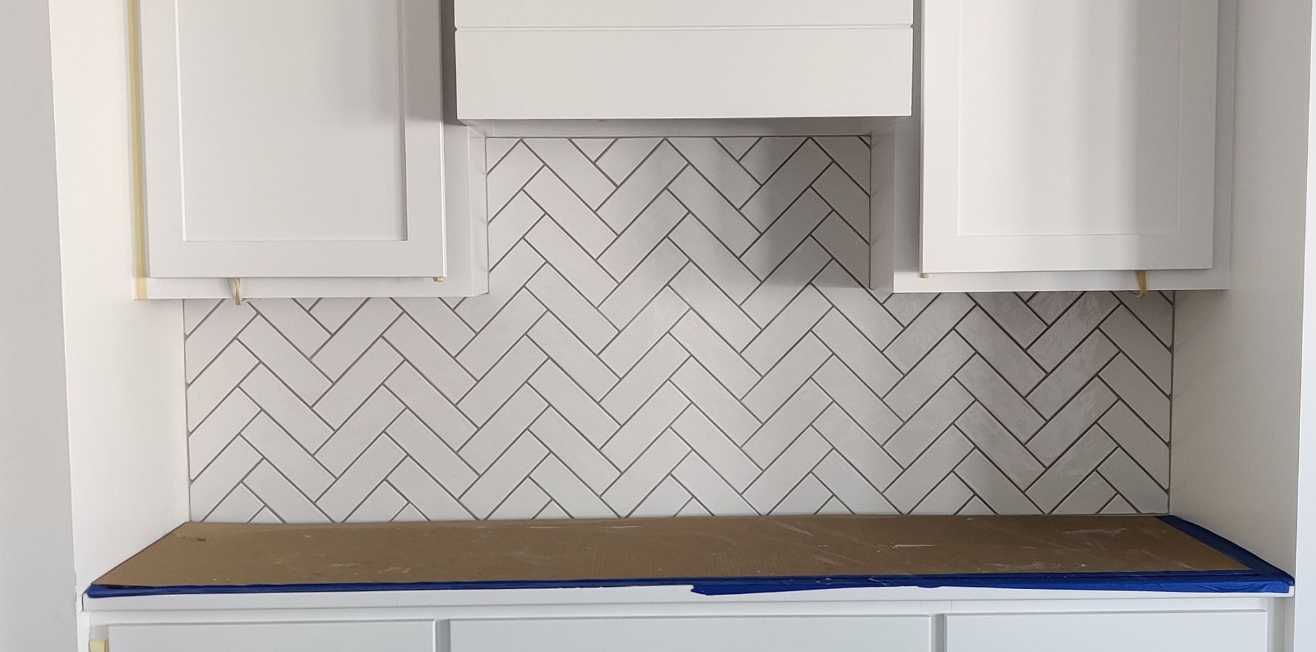 Kitchen backsplash with white herringbone tile and white cabinets above a countertop being protected with blue tape.