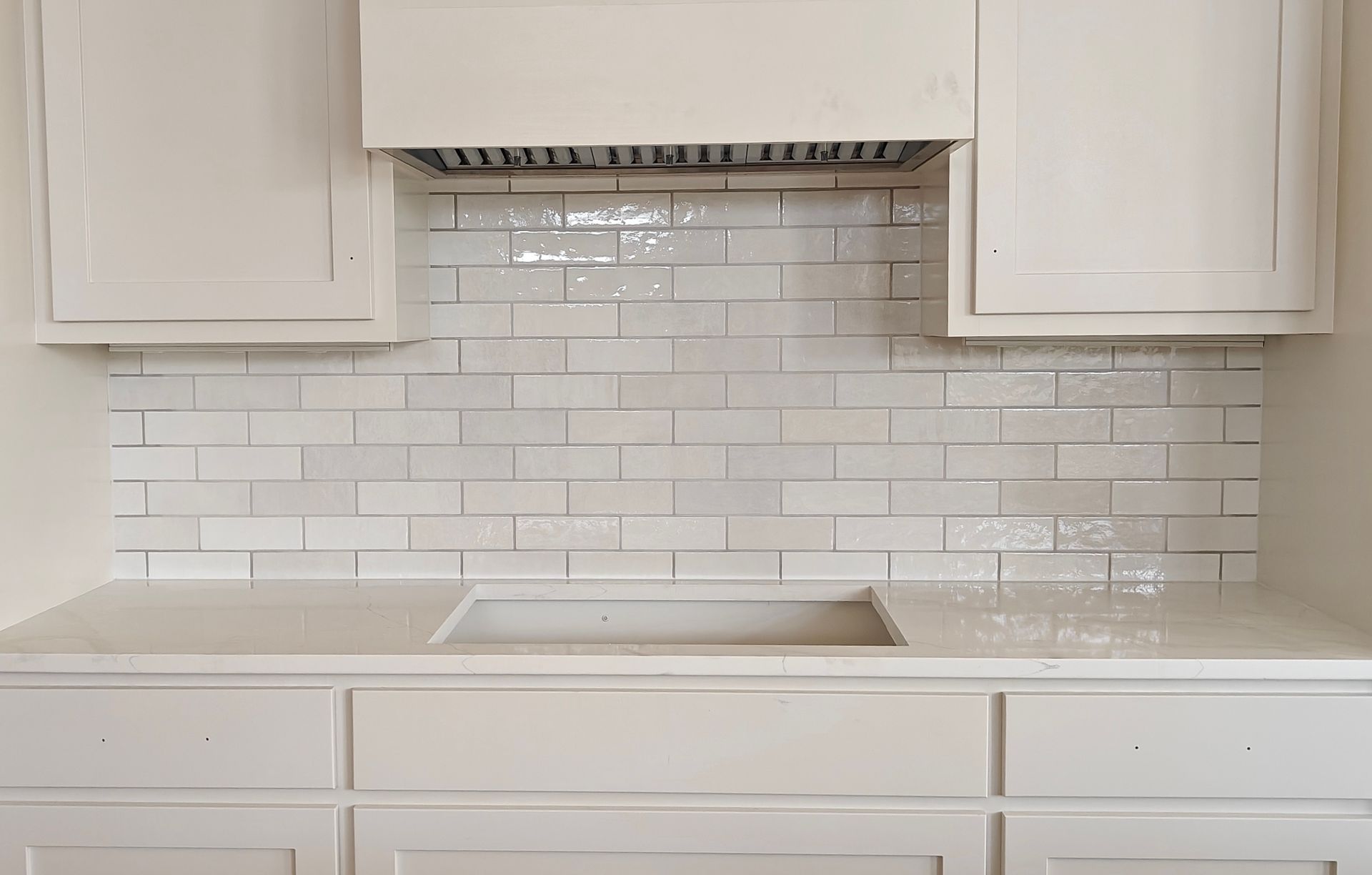 White kitchen backsplash with subway tiles, range hood, cabinets, and countertop sink area.