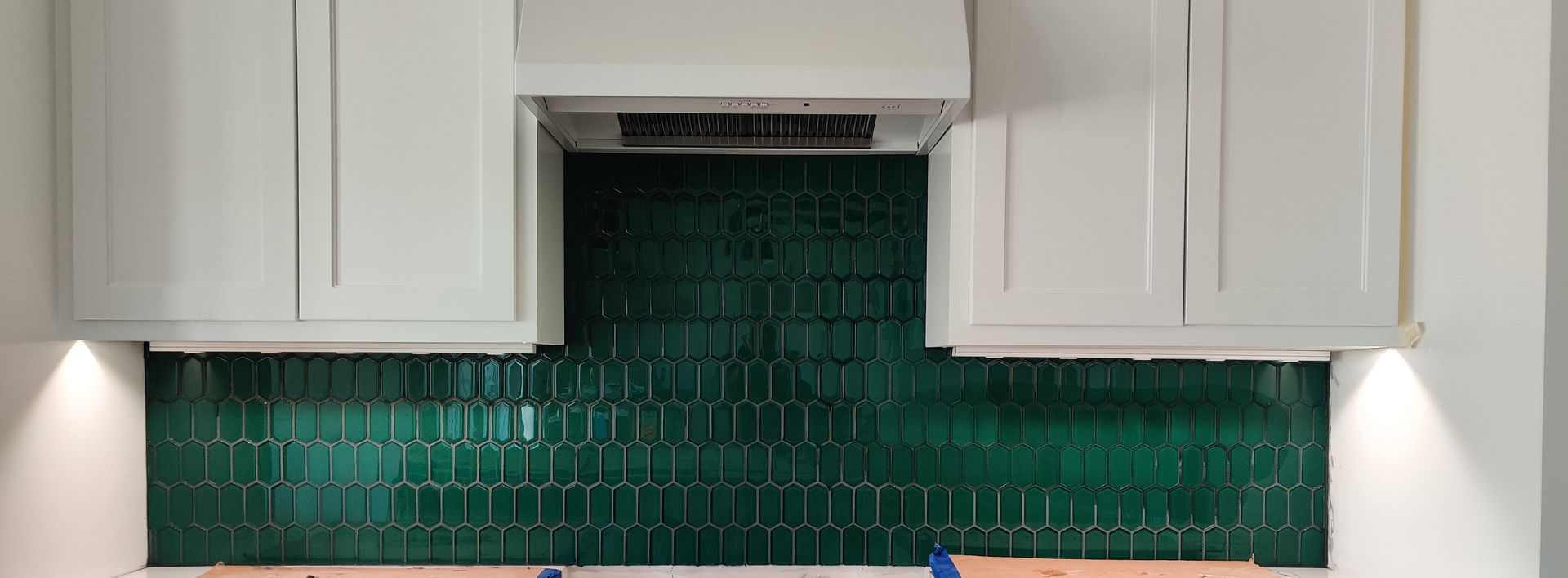 White kitchen cabinets framing a green mosaic tile backsplash under a range hood with under-cabinet lights.