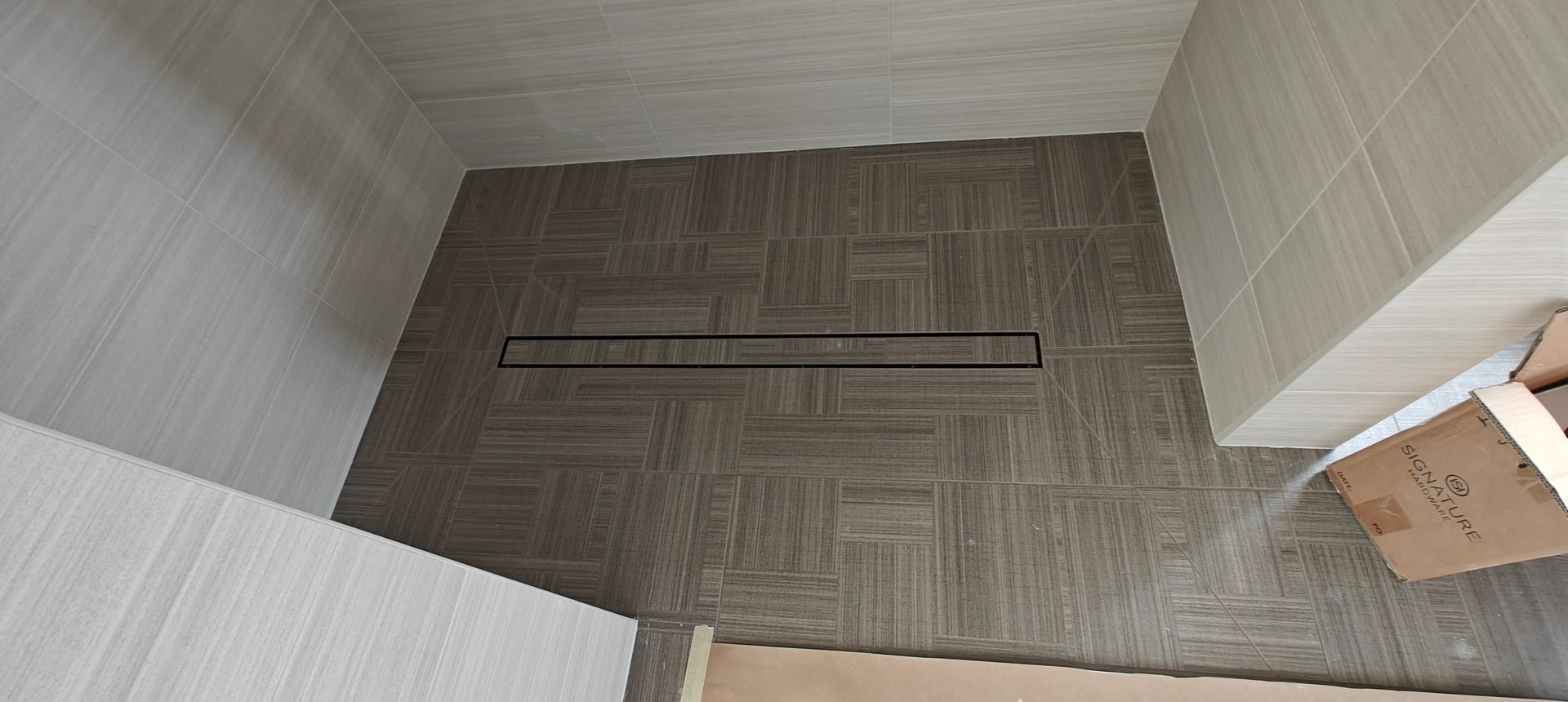 Top-down view of a small tiled corner with a long floor drain and a cardboard box on the right.