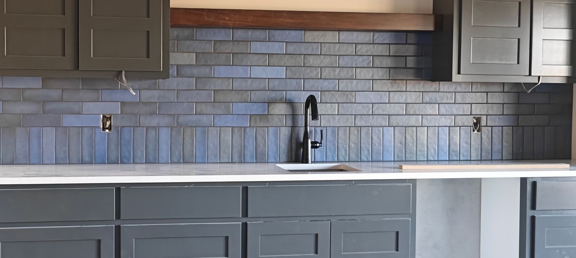 Modern kitchen with gray cabinets, white countertop, brick backsplash, and a sink under a window.