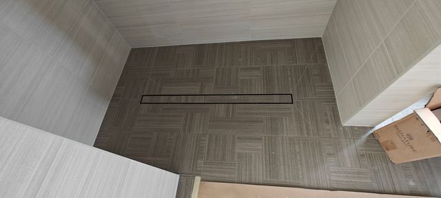 Top-down view of a tiled room corner with a long floor drain and cardboard boxes at the edge.
