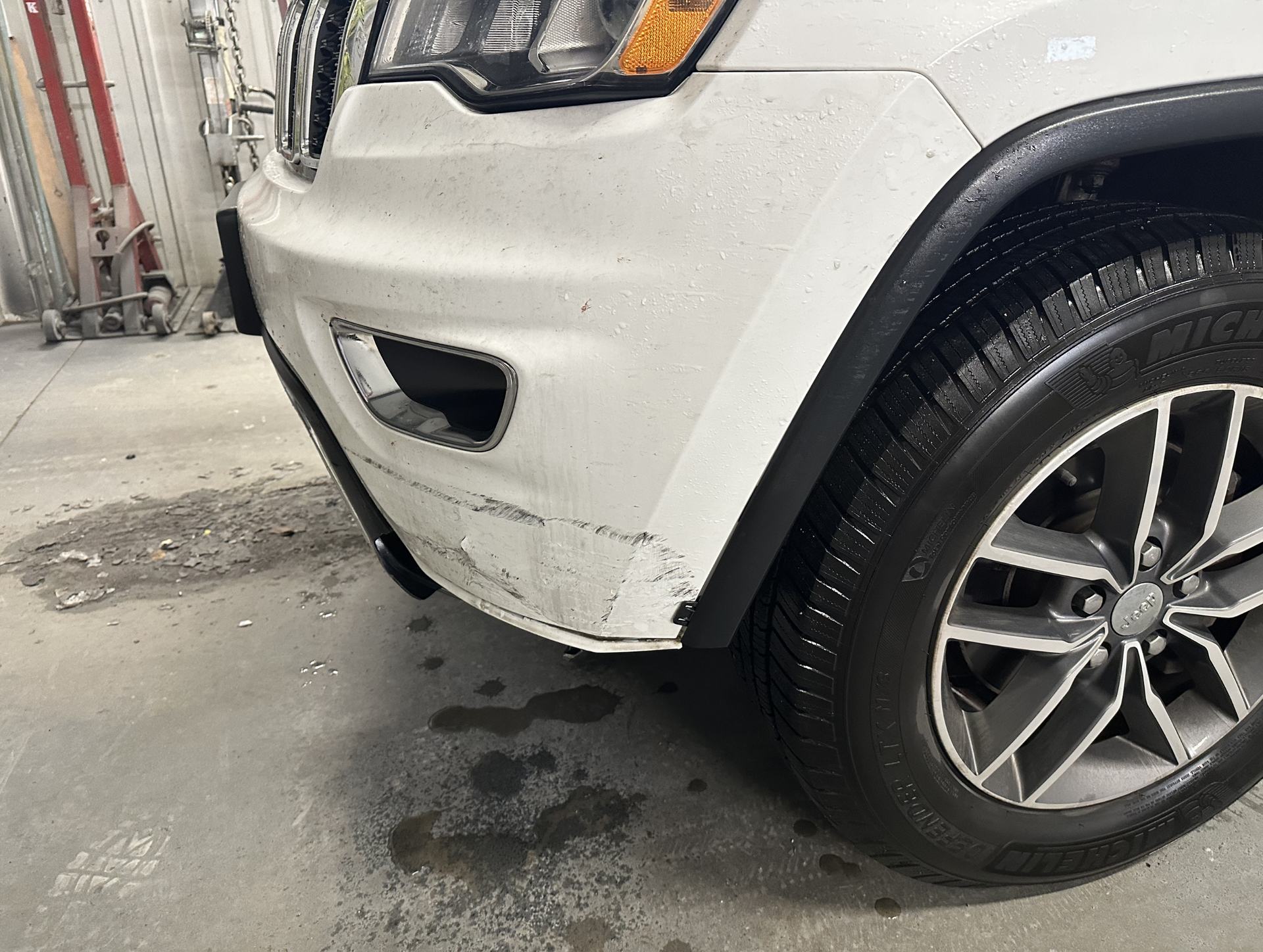White car's front bumper with scuff marks and a black fender, next to a wet tire, indoor setting.