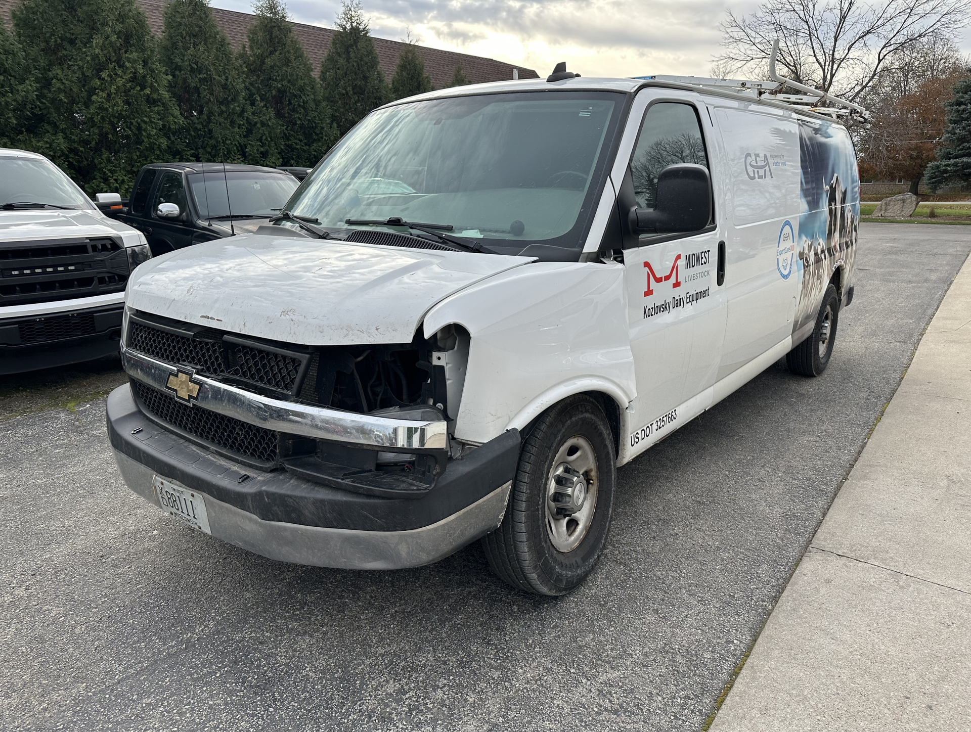 White work van with missing front grille, parked on asphalt. Logo on side, overcast sky.