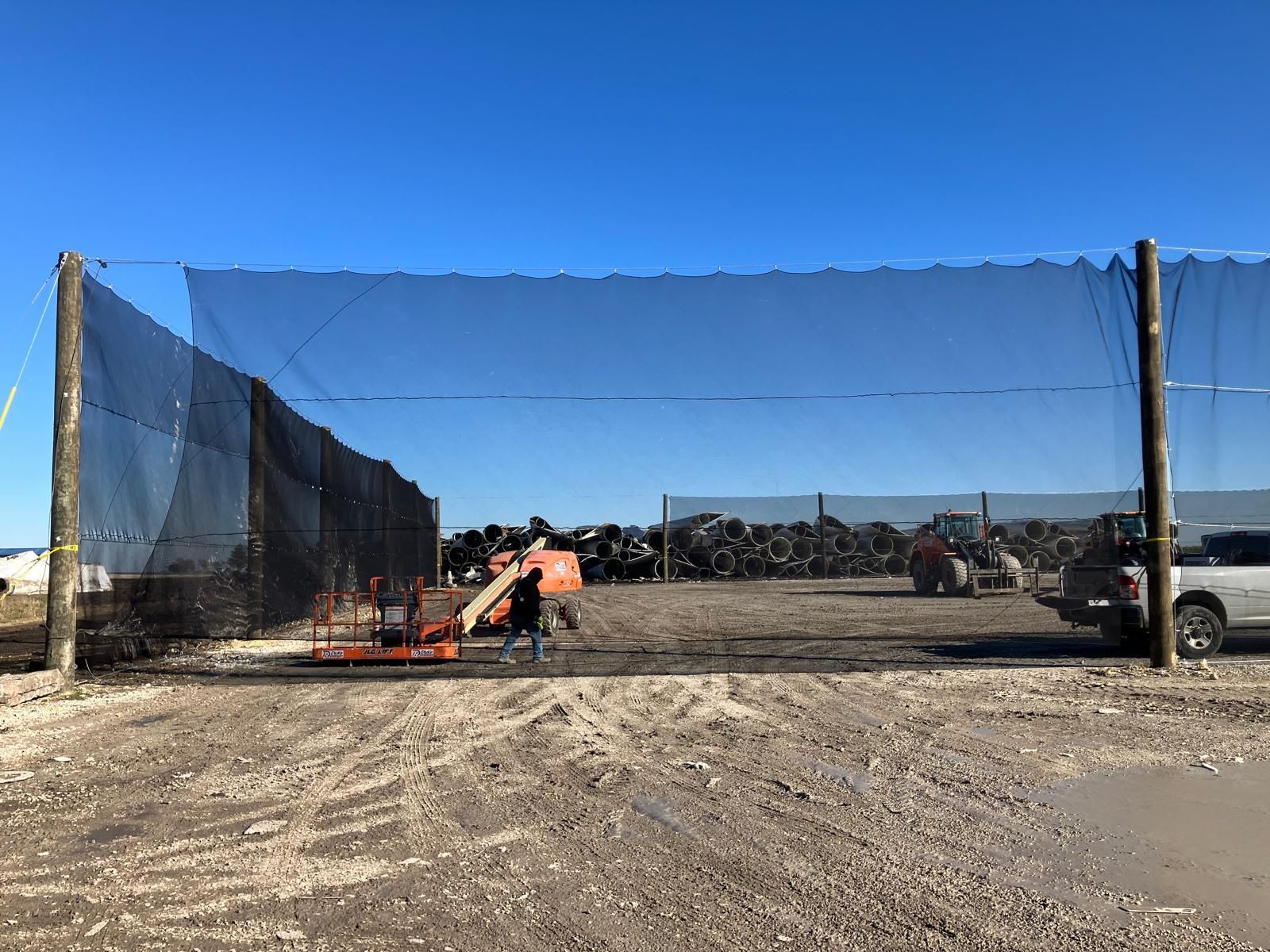 Black netting encloses a dirt lot with a forklift, vehicles, and a pile of scrap metal against a blue sky.