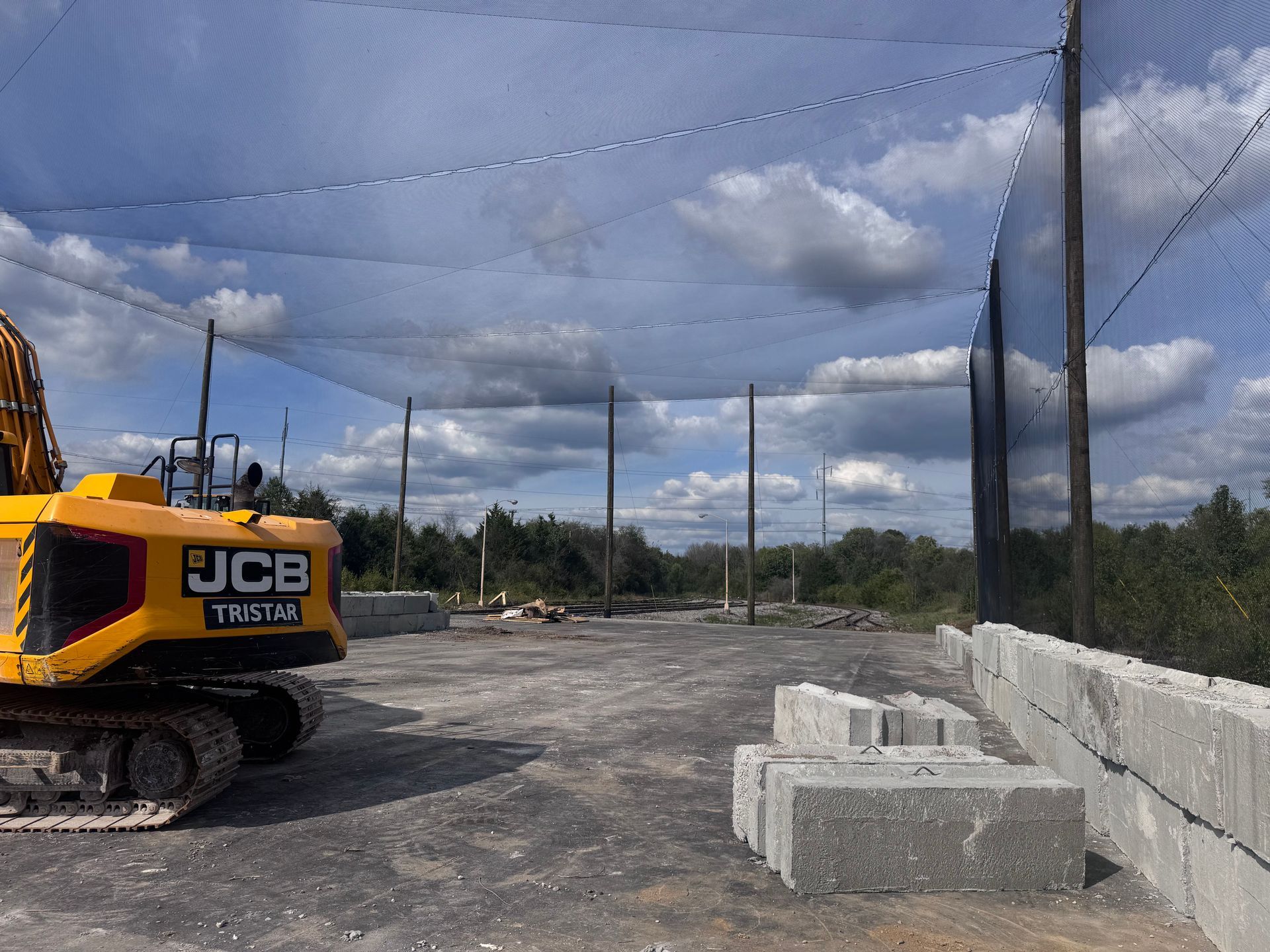 Yellow JCB excavator next to concrete blocks at construction site with netting, trees, and cloudy sky.
