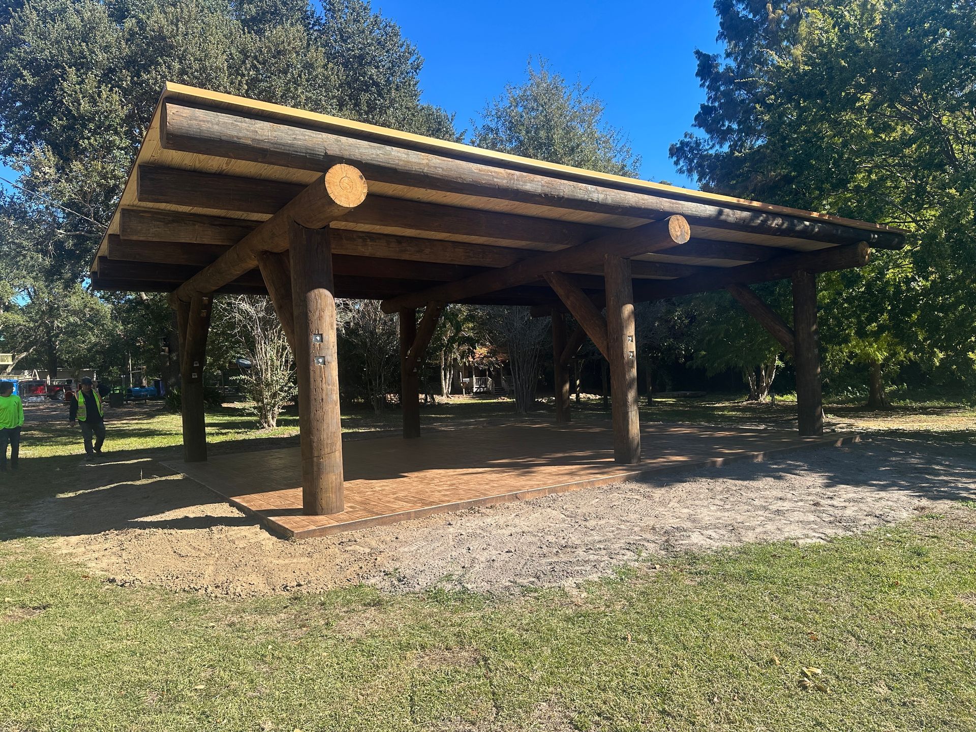 Wooden park shelter with a log roof and pillars on a gravel base. Trees and grass in the background.