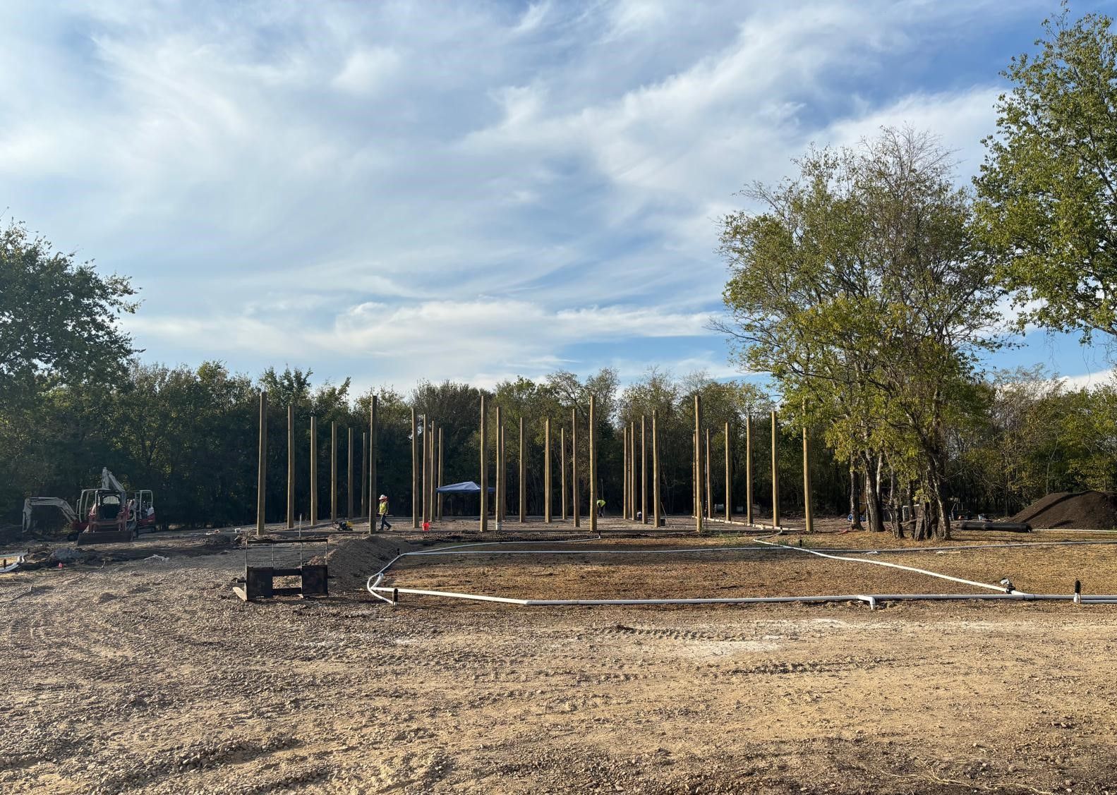 Construction site with tall wooden pillars. Clear sky with scattered clouds. Trees in background.