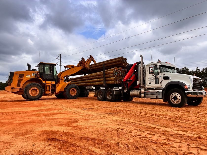 Yellow loader and white log truck on a red dirt road under cloudy sky
