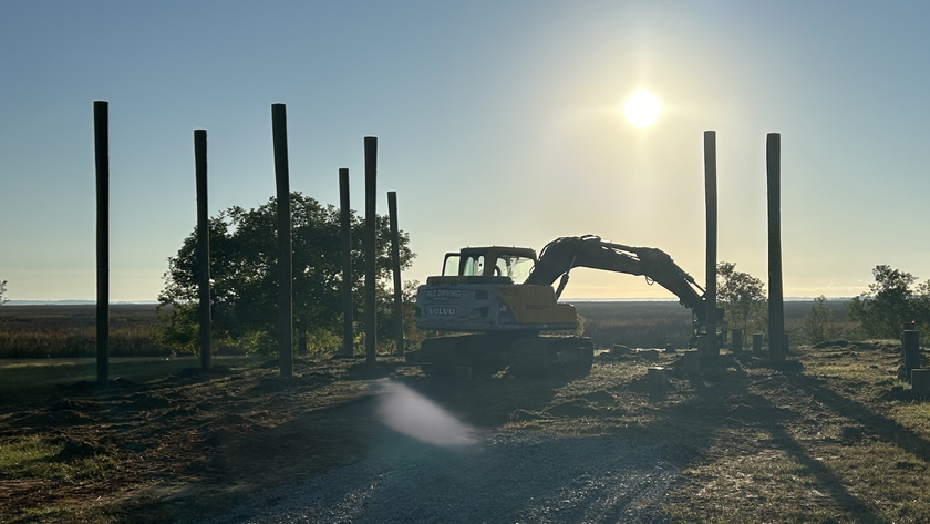 An excavator works on a construction site with wooden support poles under a bright, sunny sky.
