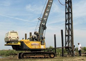Yellow construction machine with tall arm driving pilings into the ground; a person stands nearby.