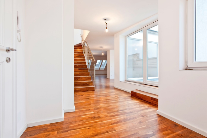 Bright empty hallway with wooden floors, stairs, and large windows overlooking a bright room
