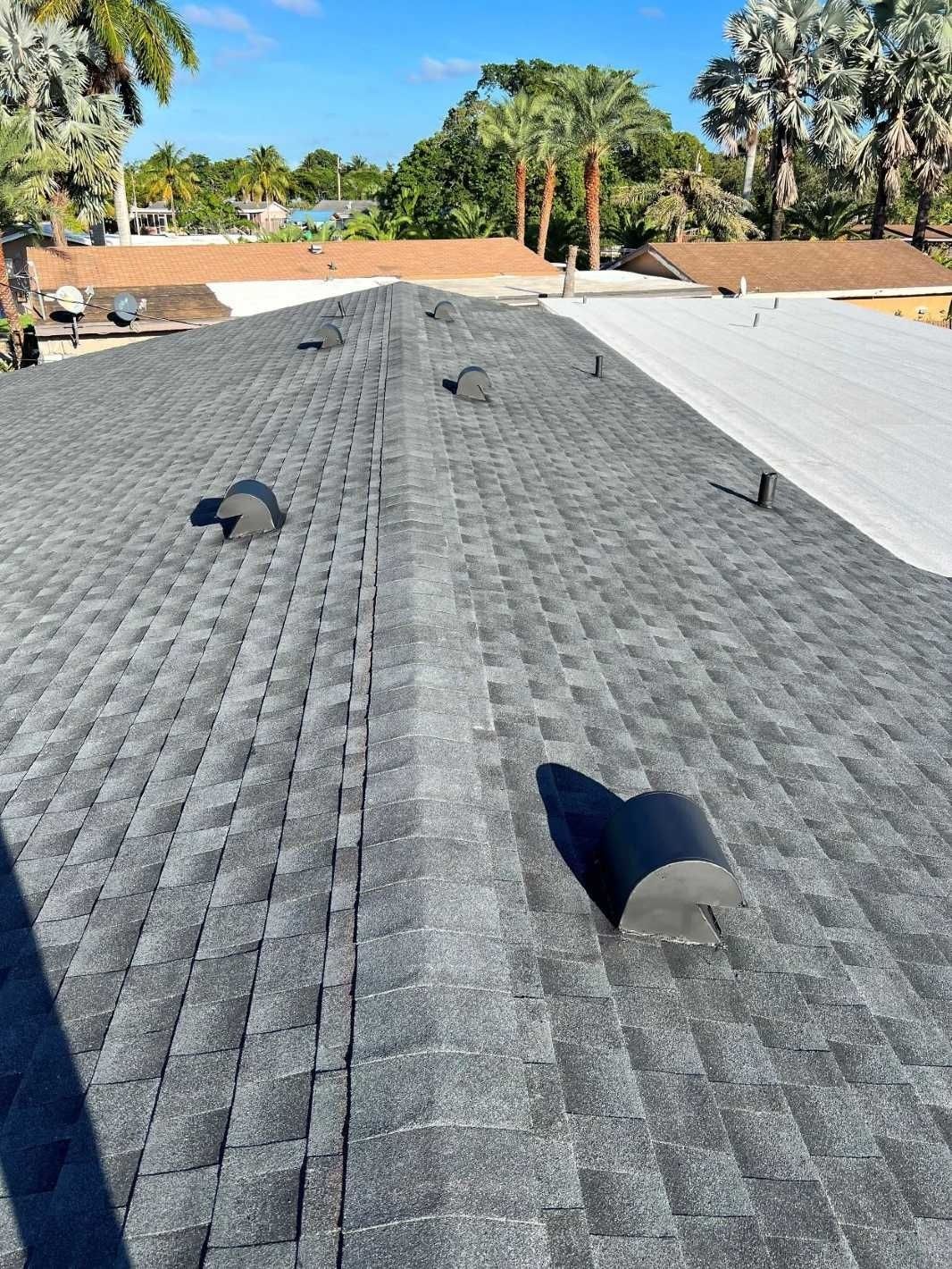 Roof with gray shingles and several vents under a clear sky, with trees and neighboring roofs in the background