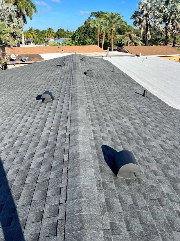Roof with gray shingles and several vents under a clear sky, with trees and neighboring roofs in the background