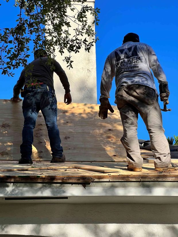 Two workers on a rooftop, looking up at a tall white wall beside a blue sky.