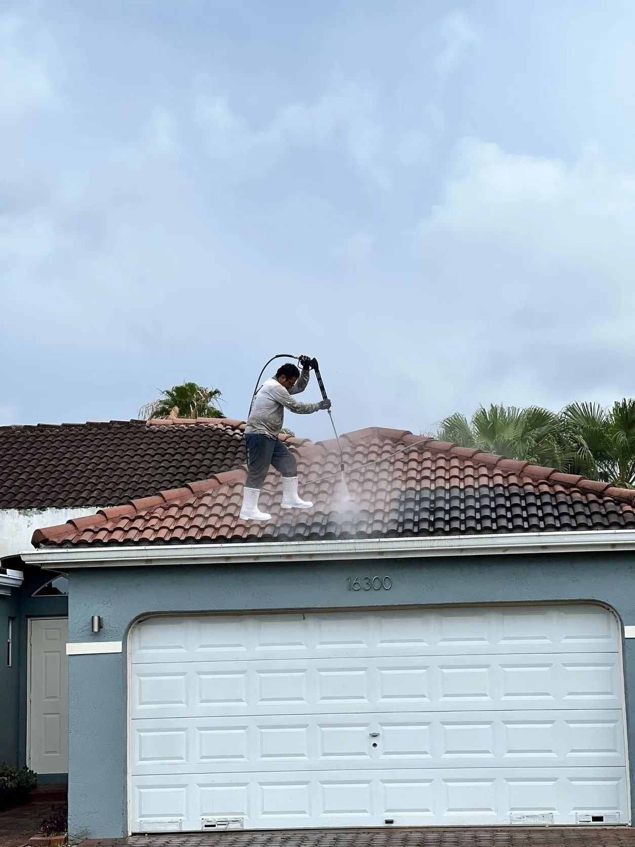 Worker power washing a red-tile roof above a white garage in a suburban home