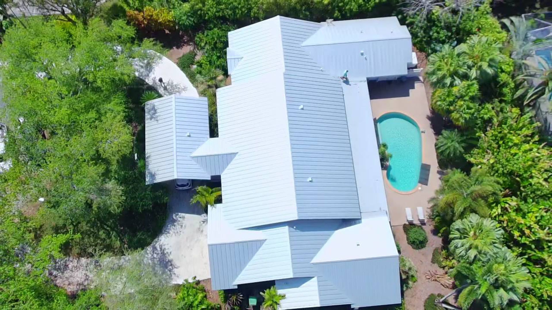 Aerial view of a white-roofed house surrounded by dense greenery, with a turquoise swimming pool in the backyard.
