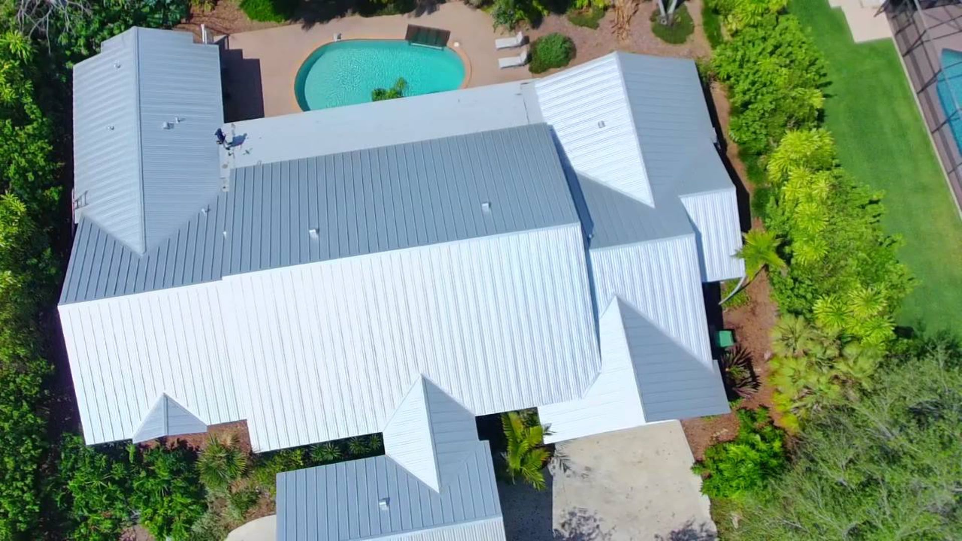Aerial view of a large white-roofed house with a swimming pool and surrounding trees.