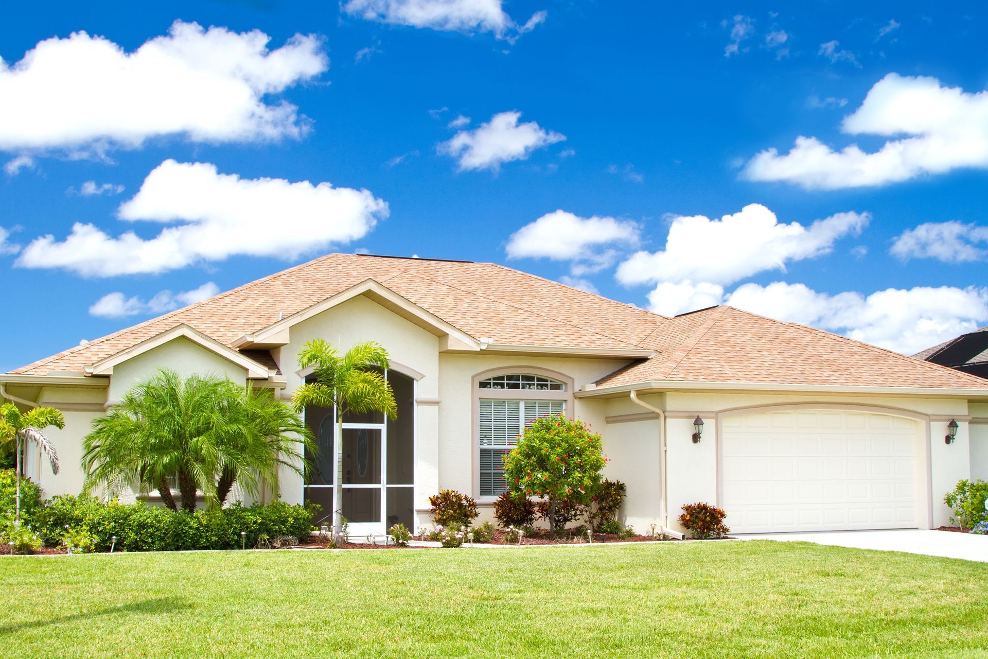 Suburban house with a tan roof, white exterior, palm trees, and a bright green lawn under a blue sky