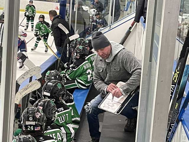 Hockey coach in gray jacket, drawing on clipboard in rink, players in green and black uniforms on bench.