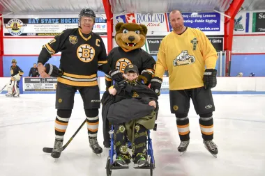 Two hockey players, a mascot, and a child in a wheelchair on an ice rink, all wearing Bruins jerseys.
