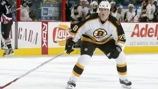 Hockey player in a black and gold Bruins jersey, skating on ice. He has a stick, in front of an advertisement.