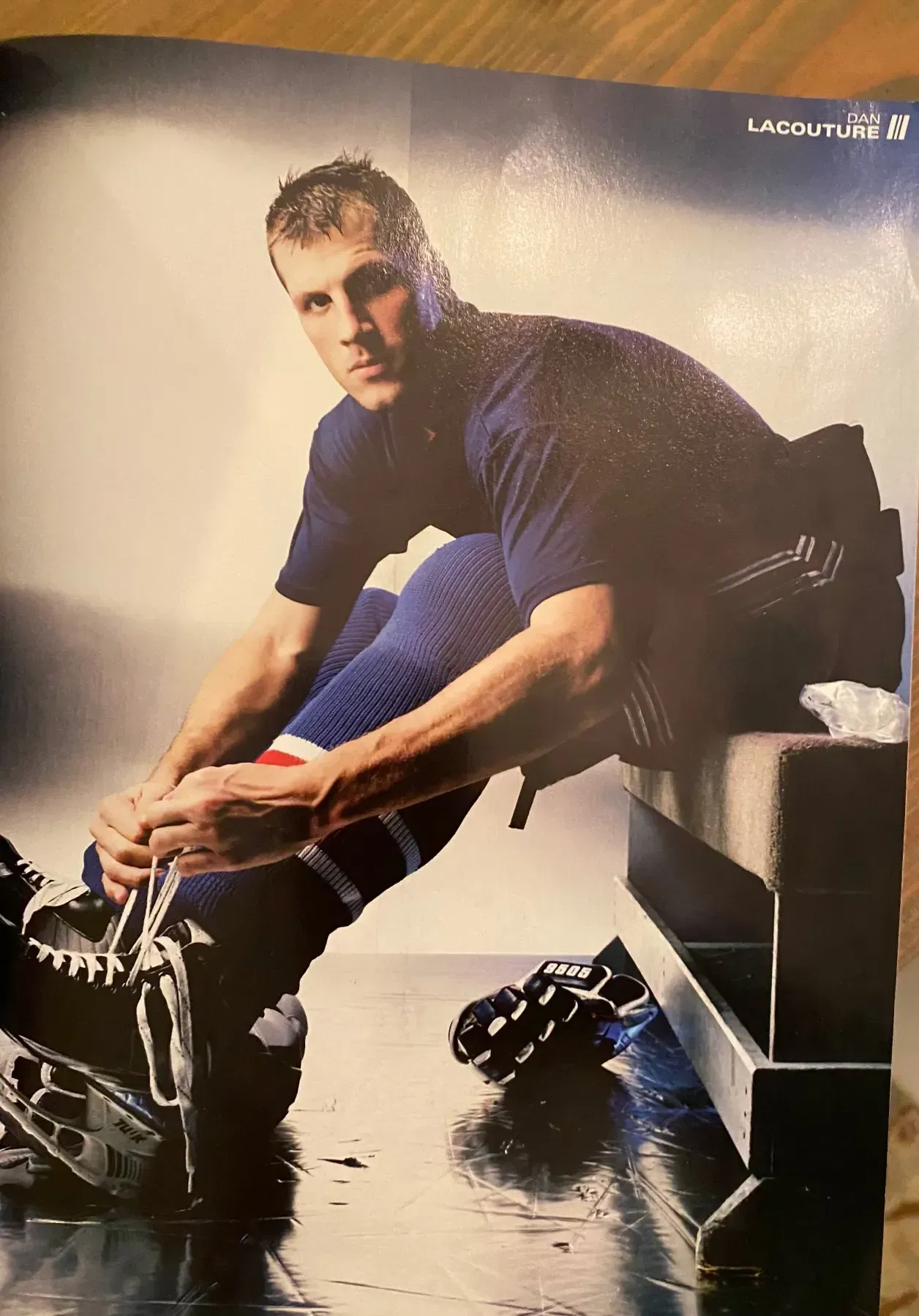 Man in hockey uniform tying his skates, seated on bench. Dark clothing, blue and red accents.