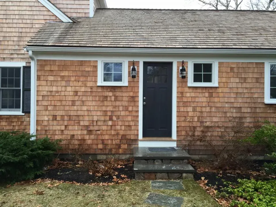 A house with brown shingles, a black door, small white windows, and a stone walkway.