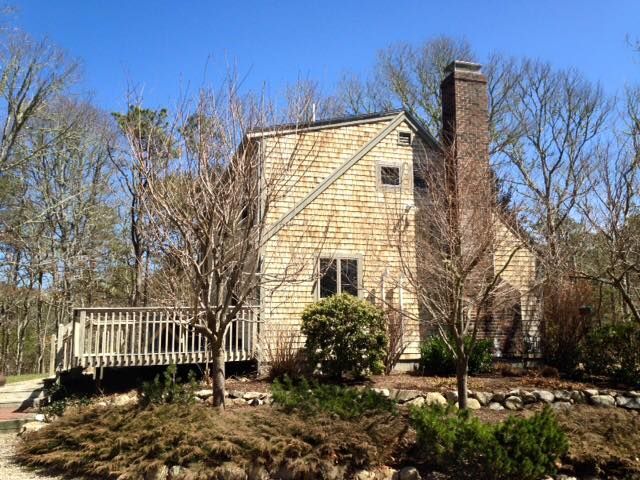 House with wood shingle siding, chimney, and wooden deck surrounded by trees and shrubs under a blue sky.