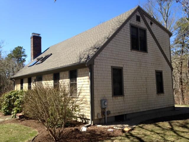 Tan house with gabled roof and chimney, set on a lawn with bushes and trees.