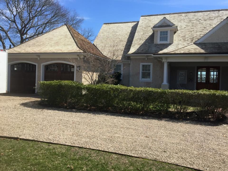 Beige house with attached garage, shingled roof, gravel driveway, and hedge.