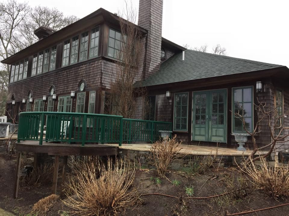 Wood-shingled house with green trim, deck, and windows. Brown bushes in front, grey sky above.