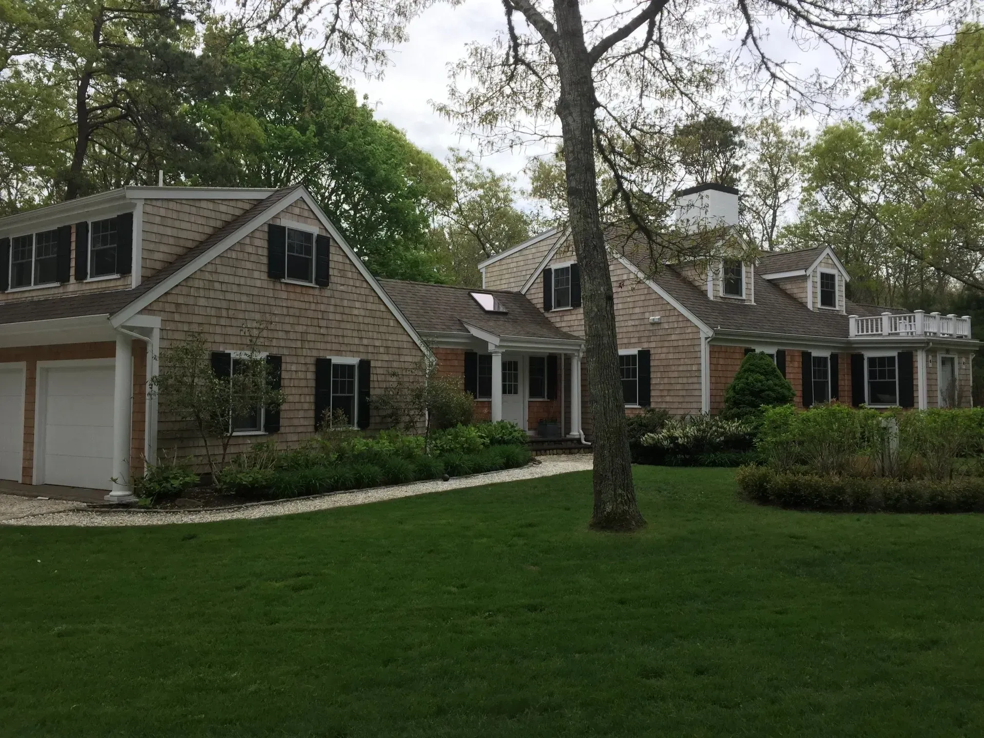 Beige brick house with black shutters, white garage doors, green lawn, and trees.