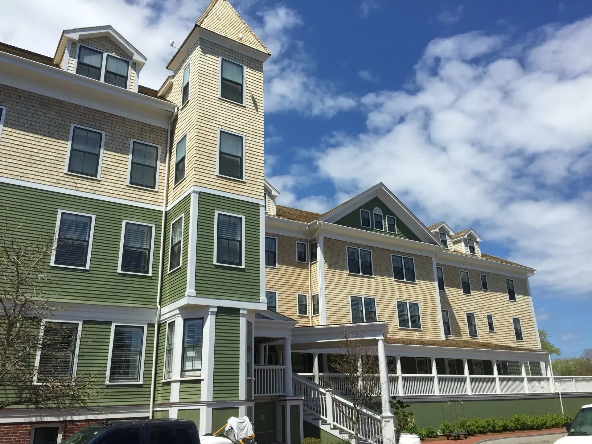Multi-story building with green and beige siding, tower, and porch under a partly cloudy sky.