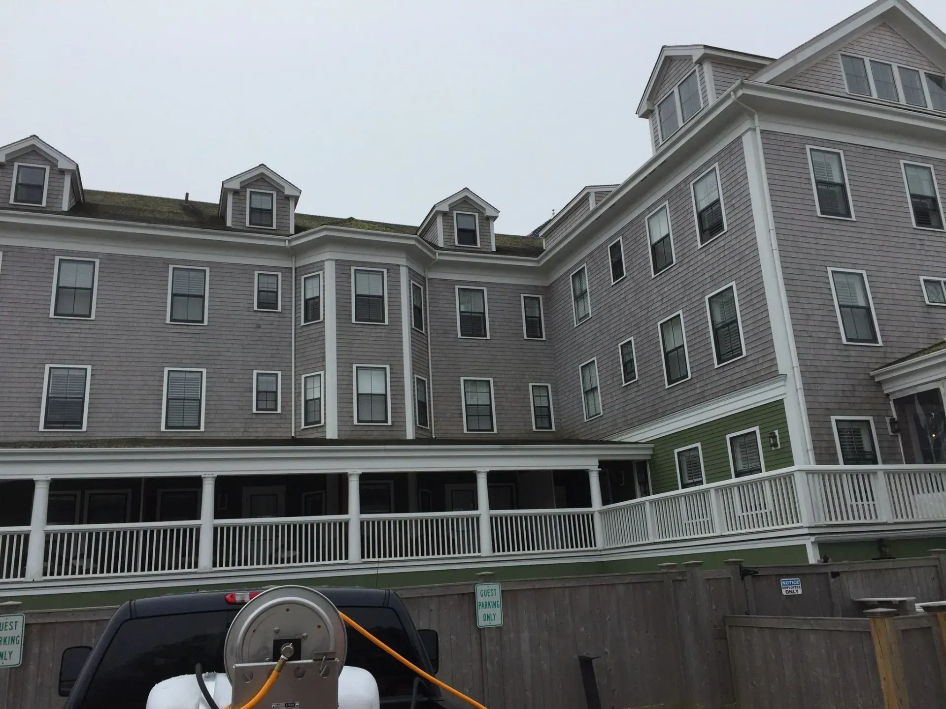Gray building with many windows and a wraparound porch, set against an overcast sky.