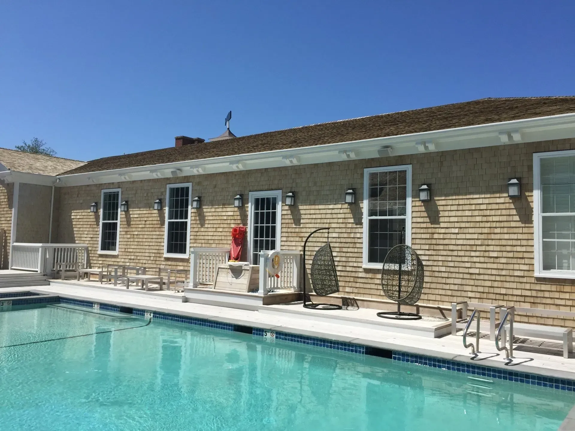 Swimming pool next to a building with windows, a brick facade, and hanging chairs; blue sky above.