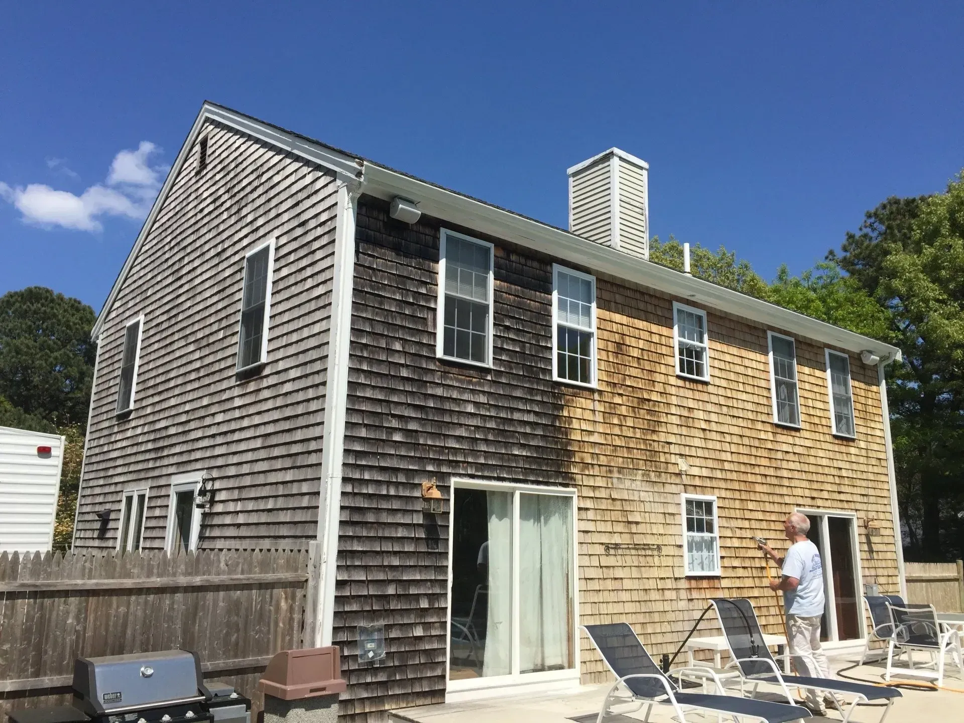 House with weathered shingle siding, half cleaned. Man stands near patio furniture, blue sky overhead.
