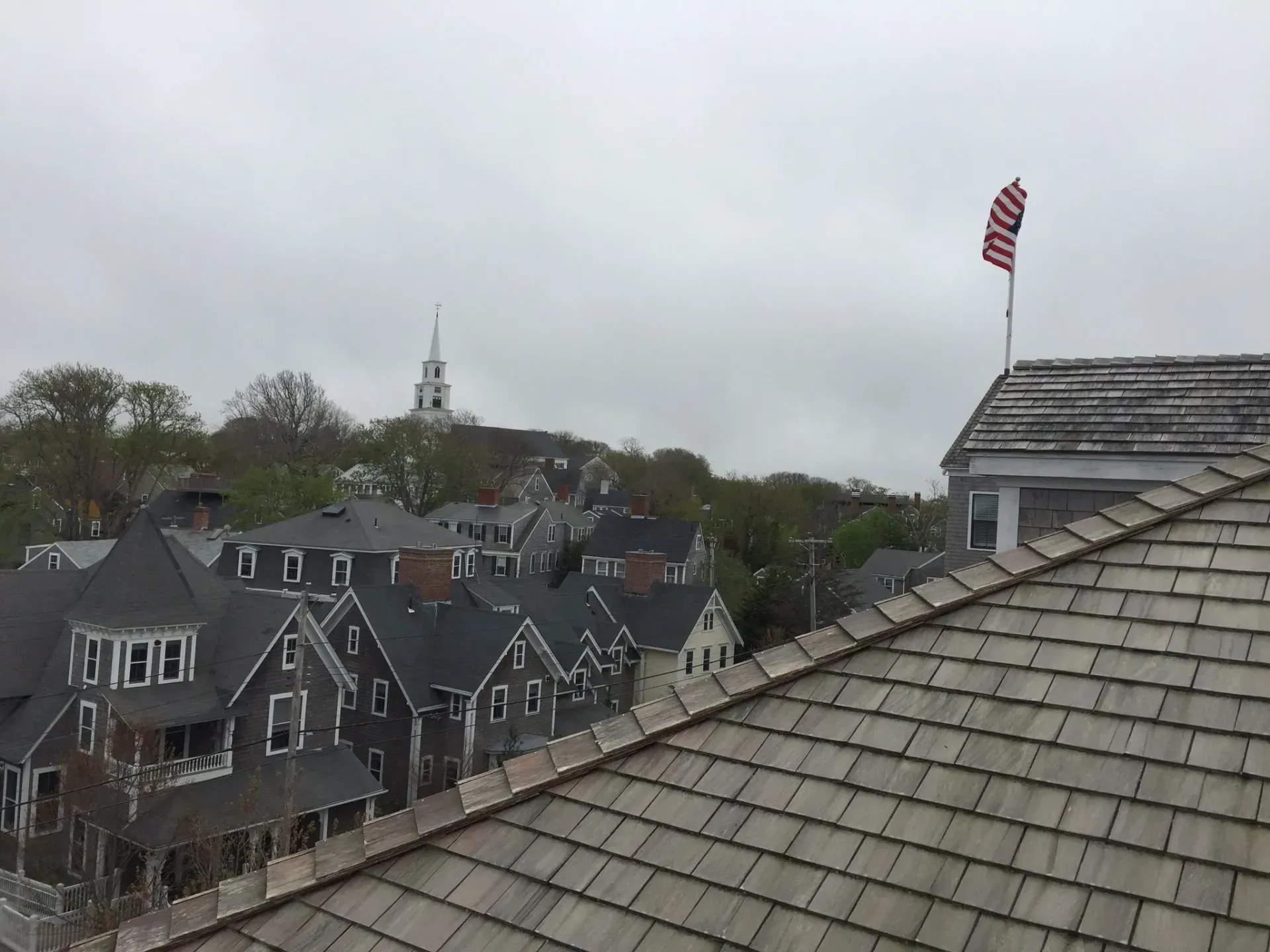 Rooftop view of gray shingled buildings, American flag, and church steeple on a cloudy day.