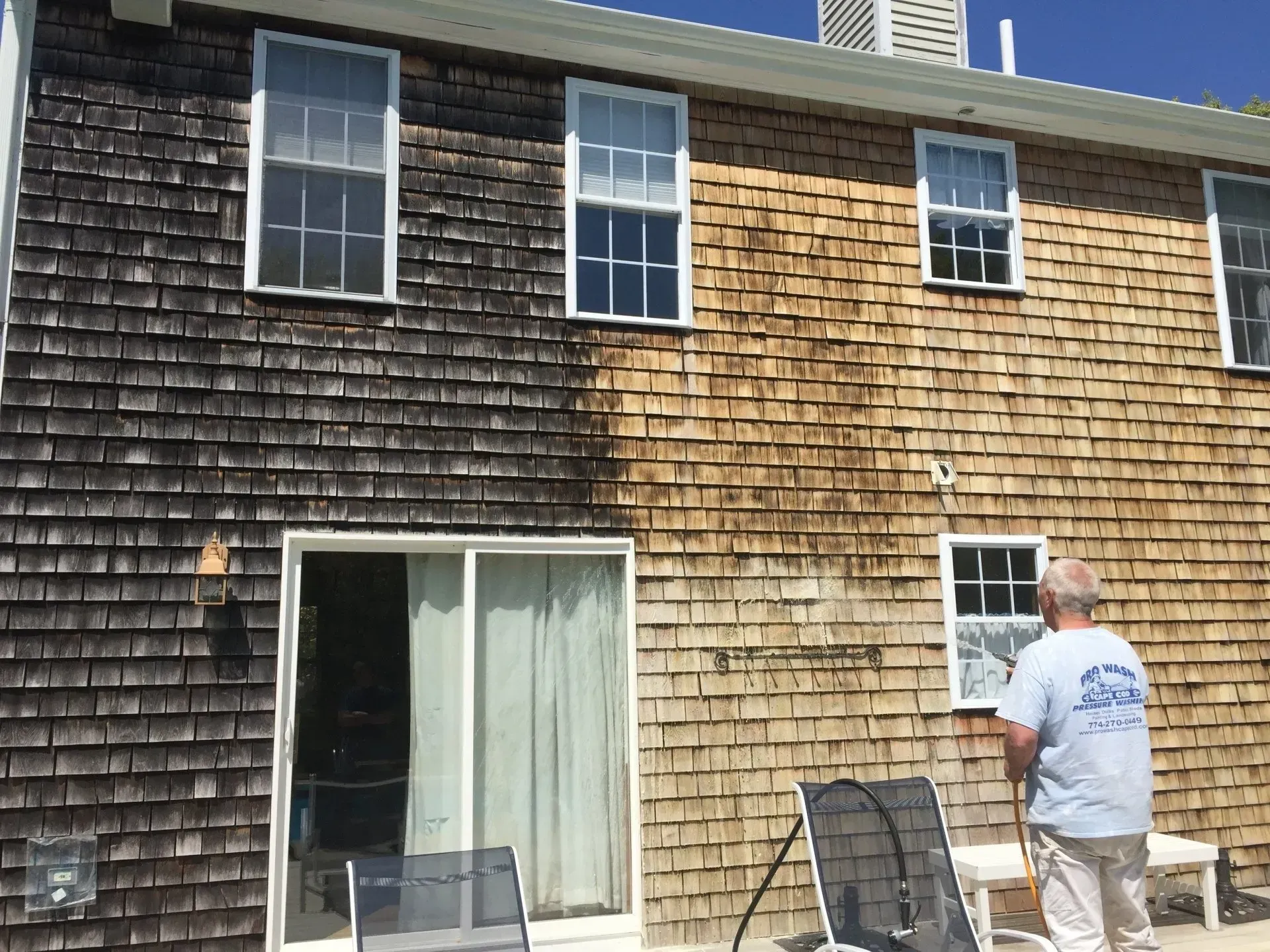 House siding being cleaned, showing a stark contrast between a dirty, dark left side and a clean, light right side. Man with hose.
