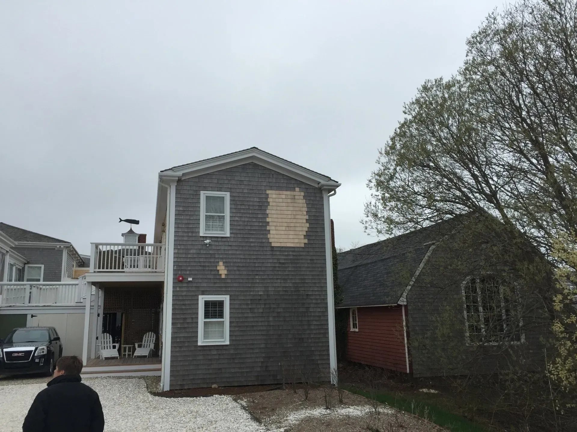 A two-story gray shingled house with a deck next to a smaller building; overcast sky.