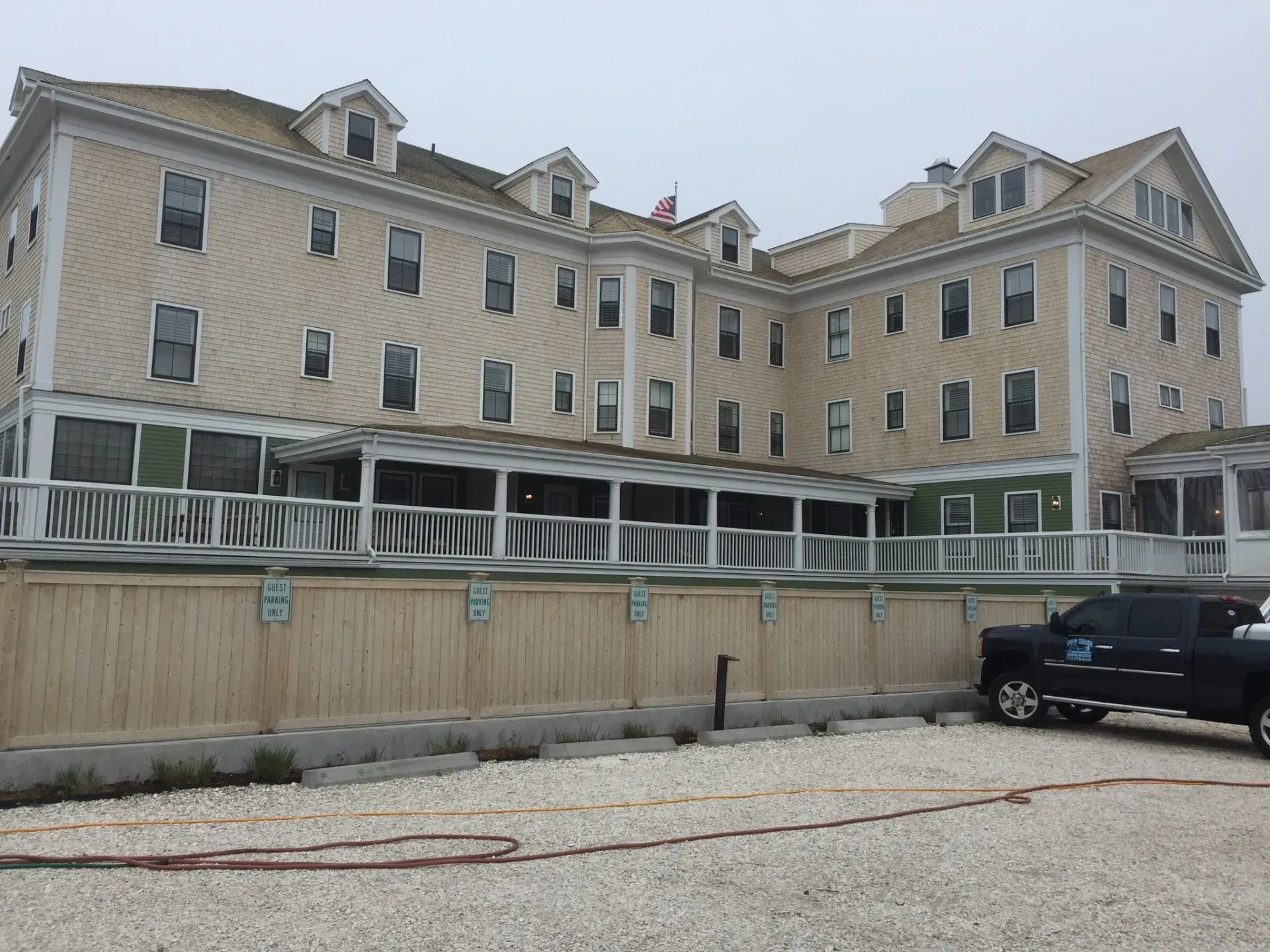 Three-story beige building with a wraparound porch, wooden fence, and black pickup truck parked in front.
