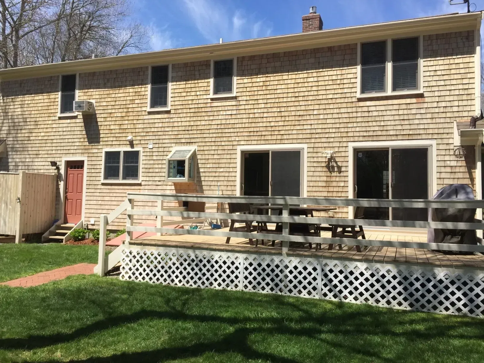 Back of a two-story beige shingled house with a deck and backyard. Red door and sliding glass doors are visible.