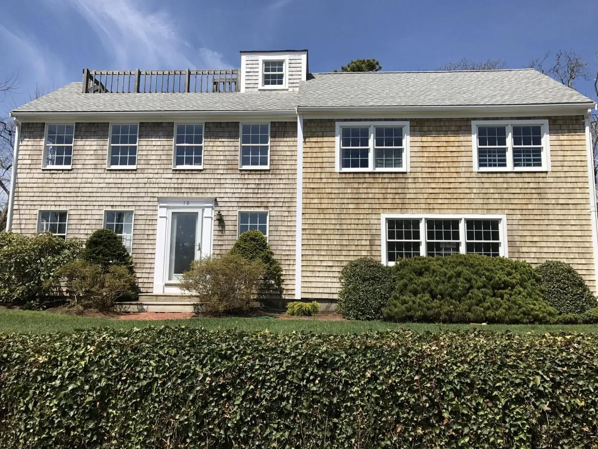Two-story house with weathered cedar shingles, multiple windows, and a small front yard with bushes.
