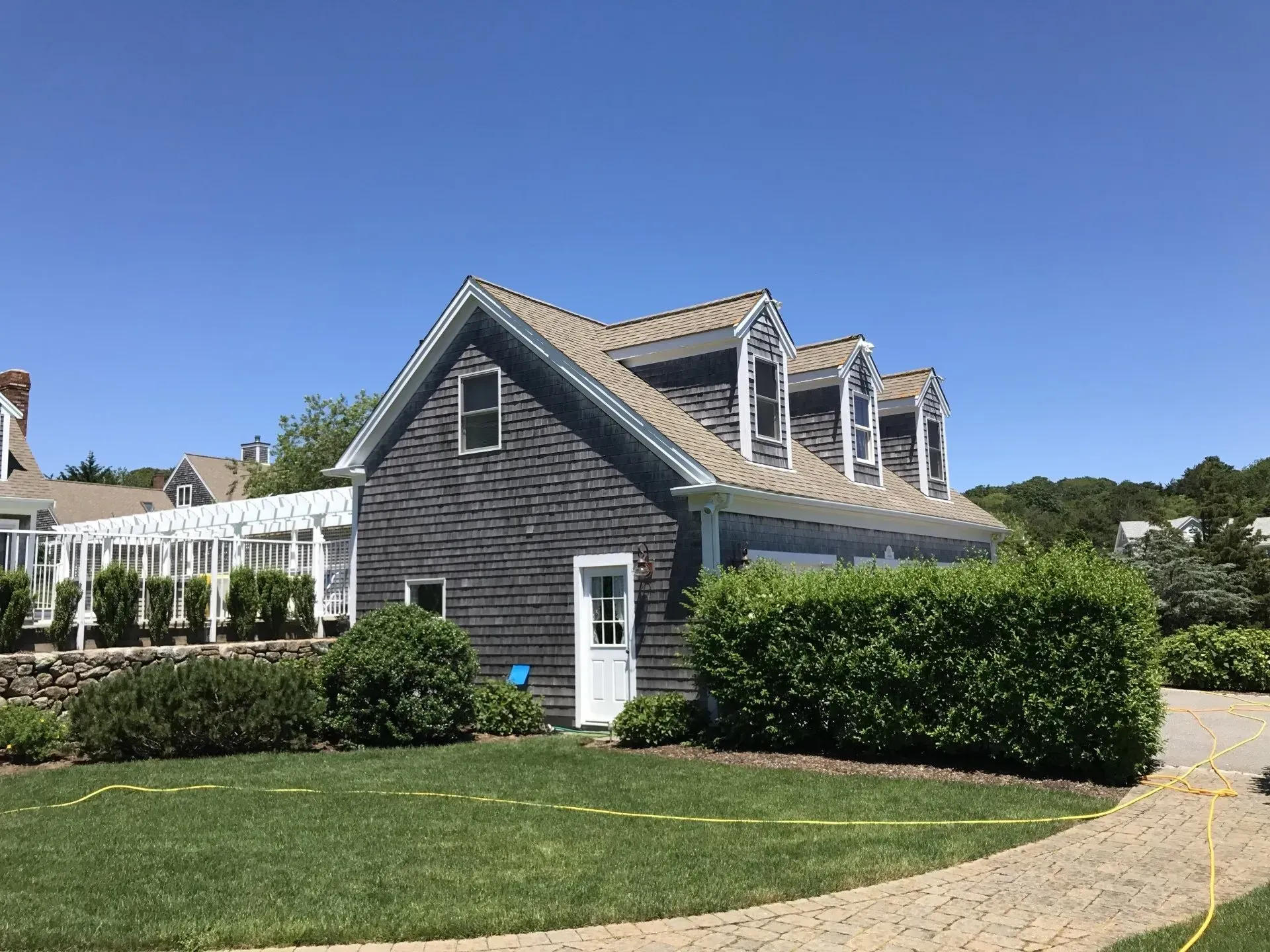Gray shingled building with dormer windows, white door, and green hedges on a sunny day.